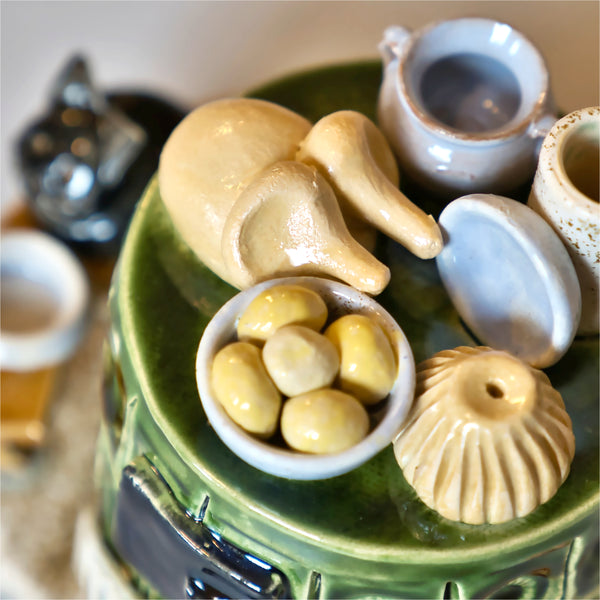 Der Keramik-Räucherofen Mini rund der Keramikwerkstatt Carola Busch hat einen grünen Sockel mit Miniaturen: einem Hasen, einer Schale mit Oliven, zwei Krügen und zwei Schüsseln.
