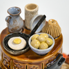 Eine Nahaufnahme von Miniatur-Keramikküchenartikeln der Keramikwerkstatt Carola Busch, darunter zwei Krüge, eine Gugelhupfform, eine Schüssel mit kleinen gelben Kugeln und eine Bratpfanne mit einem Ei. Diese bezaubernden Stücke erinnern an die Handwerkskunst des Erzgebirges und ruhen anmutig auf dem Sockel des Keramik-Räucherofens Mini rund, braun.