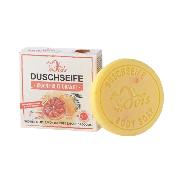 Ein rundes gelbes Stück Ovis Duschseife Grapefruit-Orange liegt neben der Schachtel, die Bilder von Grapefruit und Orange zeigt und den Duft als Grapefruit-Orange kennzeichnet.