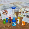 Auf einem Spielbrett sind bunte Figuren und Würfel neben Karten angeordnet, die auf ein Fantasy-Abenteuer hinweisen. Eine blaue Sanduhr ergänzt die Szene von „Abenteuer im Liederwald“ Edition Kinderlieder der Brettschneider Verpackungen GmbH, angesiedelt im erzgebirgischen Liederwald.