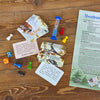 Auf einem Holztisch liegt das Brettspiel „Abenteuer im Liederwald“ Edition Kinderlieder der Brettschneider Verpackungen GmbH mit zwei Karten mit der Aufschrift „Aktion“ und „Ereignis“, bunten Spielfiguren, einer blauen Sanduhr, Würfeln und einem Regelblatt für eine spannende Reise durch den erzgebirgischen Liederwald.