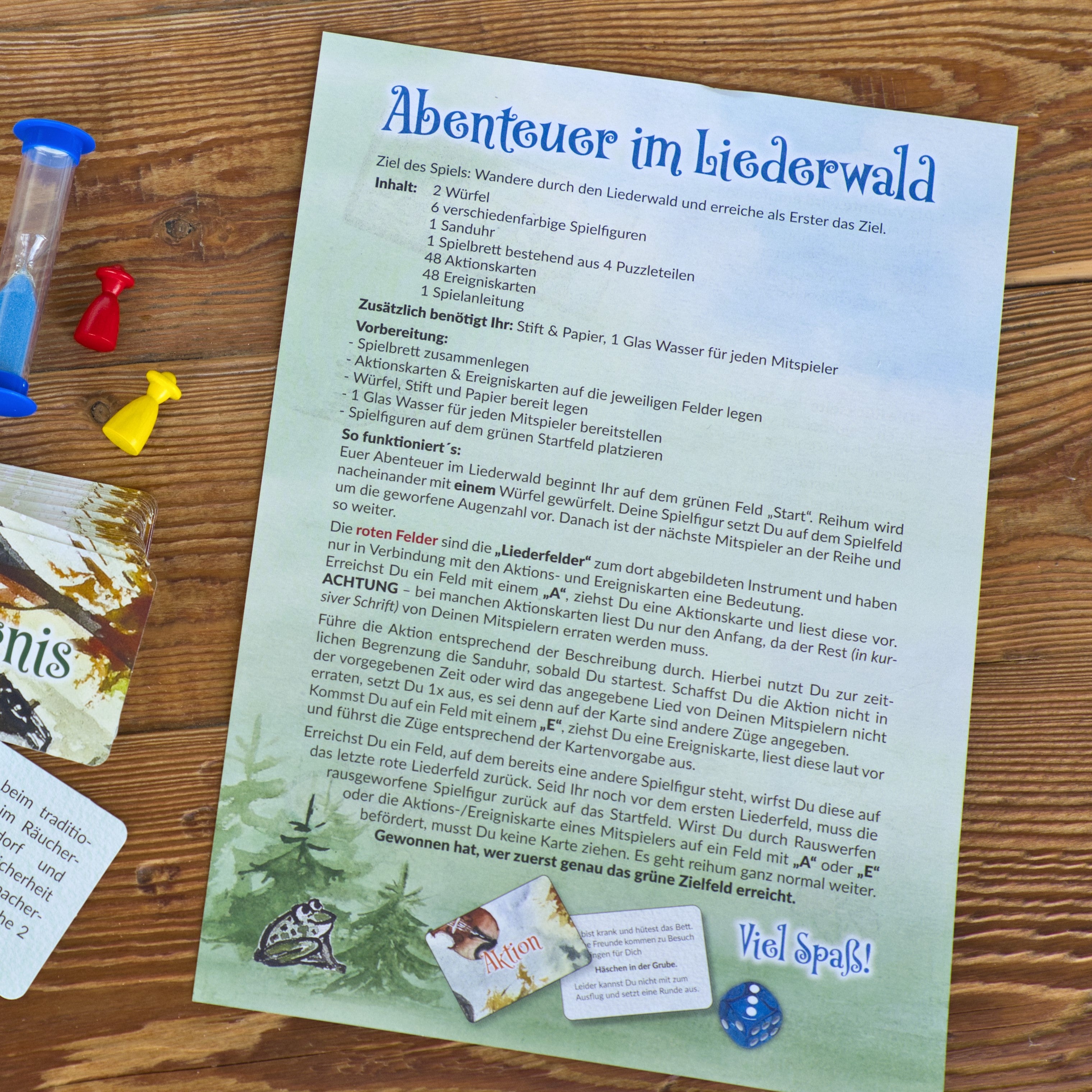 Die deutsche Spielanleitung für das Brettspiel „Abenteuer im Liederwald“ von Brettschneider Verpackungen GmbH befindet sich auf einem Holztisch mit einer Sanduhr, blauen und roten Spielbrettsteinen und einem gelben Würfel. Regeln und Ziele sind beschrieben, und auf der Seite sind teilweise Spielkarten sichtbar.