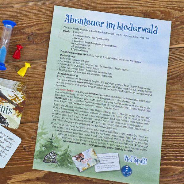 Die deutsche Spielanleitung für das Brettspiel „Abenteuer im Liederwald“ von Brettschneider Verpackungen GmbH befindet sich auf einem Holztisch mit einer Sanduhr, blauen und roten Spielbrettsteinen und einem gelben Würfel. Regeln und Ziele sind beschrieben, und auf der Seite sind teilweise Spielkarten sichtbar.