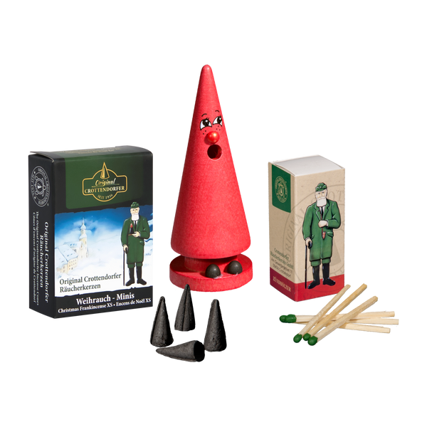 Eine rote kegelförmige Holzfigur mit Gesicht steht neben einem Mini-Räucherset „Romy Rose“ und einer Original Crottendorfer-Dose mit einem Mann in Grün. Im Vordergrund liegen verstreut drei Mini-Räucherkerzen und mehrere Streichhölzer.