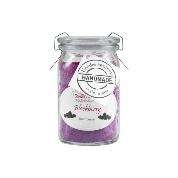 Eine Candle Factory „Blackberry“ Duftkerze/Windlicht im Glasgefäß, mit sattem violettem pflanzlichem Stearinwachs mit Brombeer-Note, inklusive kleinem Etikett um den Hals und „Handmade in Germany“.