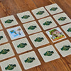 Auf einem Holztisch steht ein Spiel „Finde2“ von Brettschneider Verpackungen GmbH, mit mehreren verdeckten Karten. Zwei Karten sind aufgedeckt und zeigen unterschiedliche Illustrationen. Alle haben auf der Rückseite ein grün-goldenes „Crest & Showmen“-Logo, das diesem spannenden Bildpaare-Spiel Eleganz verleiht.