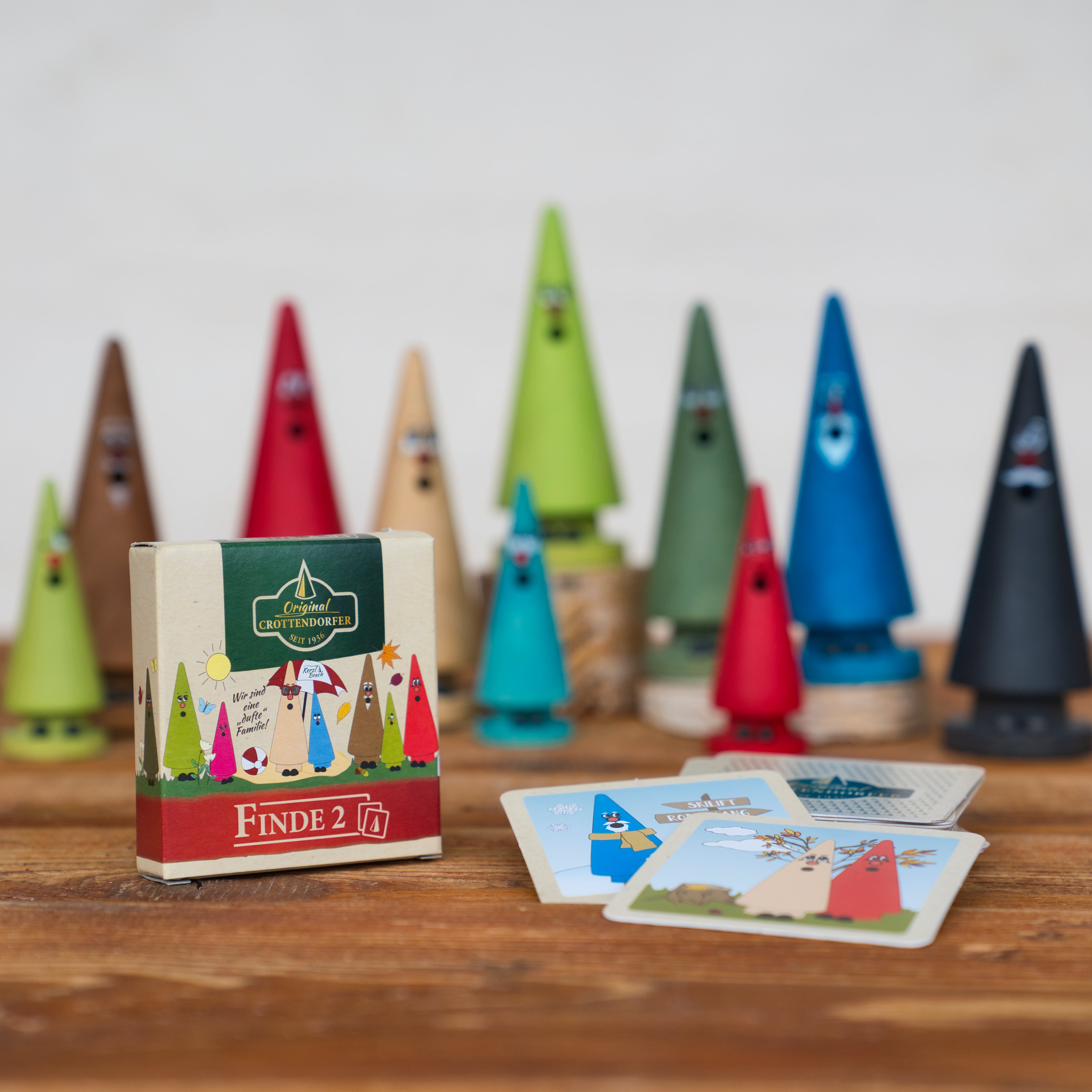 Das Spiel „Finde2“ der Brettschneider Verpackungen GmbH besteht aus kegelförmigen, bunten Baumfiguren aus Holz mit Gesichtern und kleinen Bildpaare-Karten mit ähnlichen Figuren. Auf einer Holzoberfläche platziert, erzeugen die Box und die Karten eine spielerische Memospiel-Atmosphäre.