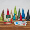 Das Spiel „Finde2“ der Brettschneider Verpackungen GmbH besteht aus kegelförmigen, bunten Baumfiguren aus Holz mit Gesichtern und kleinen Bildpaare-Karten mit ähnlichen Figuren. Auf einer Holzoberfläche platziert, erzeugen die Box und die Karten eine spielerische Memospiel-Atmosphäre.