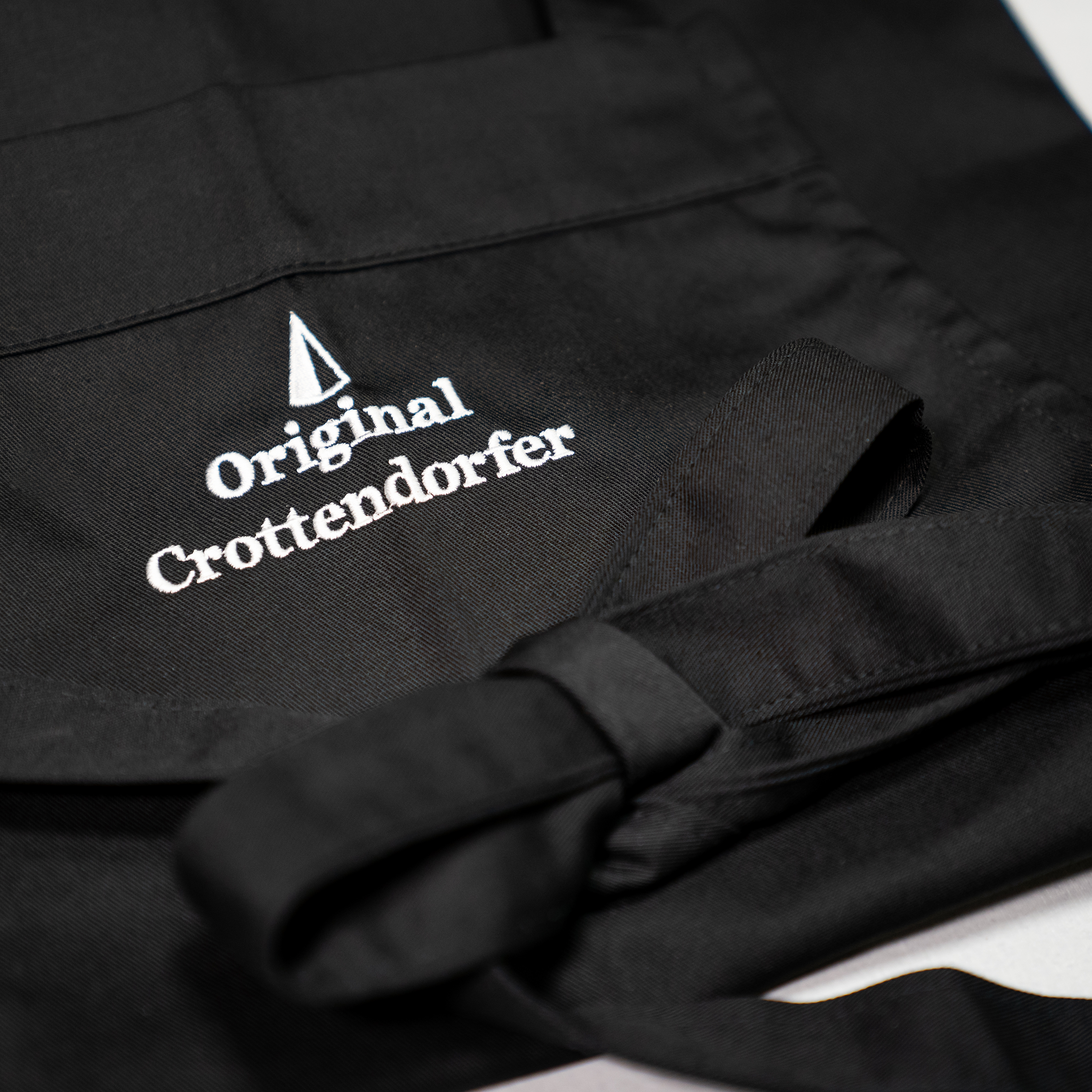 Crottendorfer Räucherkerzen-Schürze schwarz, gefaltet, mit Detailansicht vom Logo und Schleife