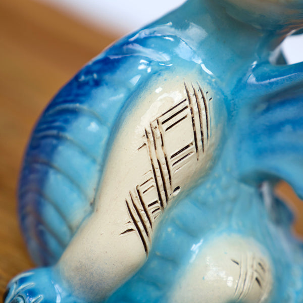 Eine Nahaufnahme der Keramikskulptur „KU Drache Blau“ von Kurilin DECO zeigt einen blau-weißen Drachen mit strukturierten Schuppen und geätzten Mustern auf einem cremefarbenen Arm. Eine unscharfe Holzoberfläche im Hintergrund ergänzt diese Keramikkunst.