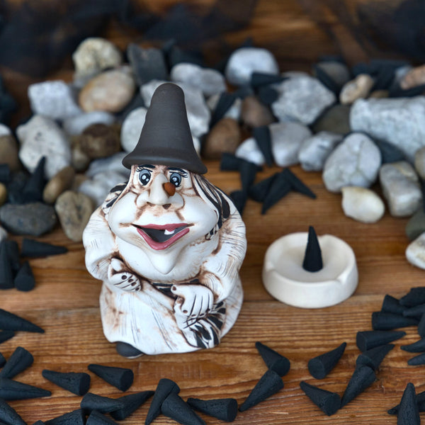 Die Keramikfigur KU Hexe „Baba Jaga“ von Kurilin DECO trägt einen braunen Spitzhut und ist von kleinen schwarzen kegelförmigen Räucherkerzen umgeben, von denen einige in einer Schale daneben liegen. Verstreute Steine bilden den Hintergrund.