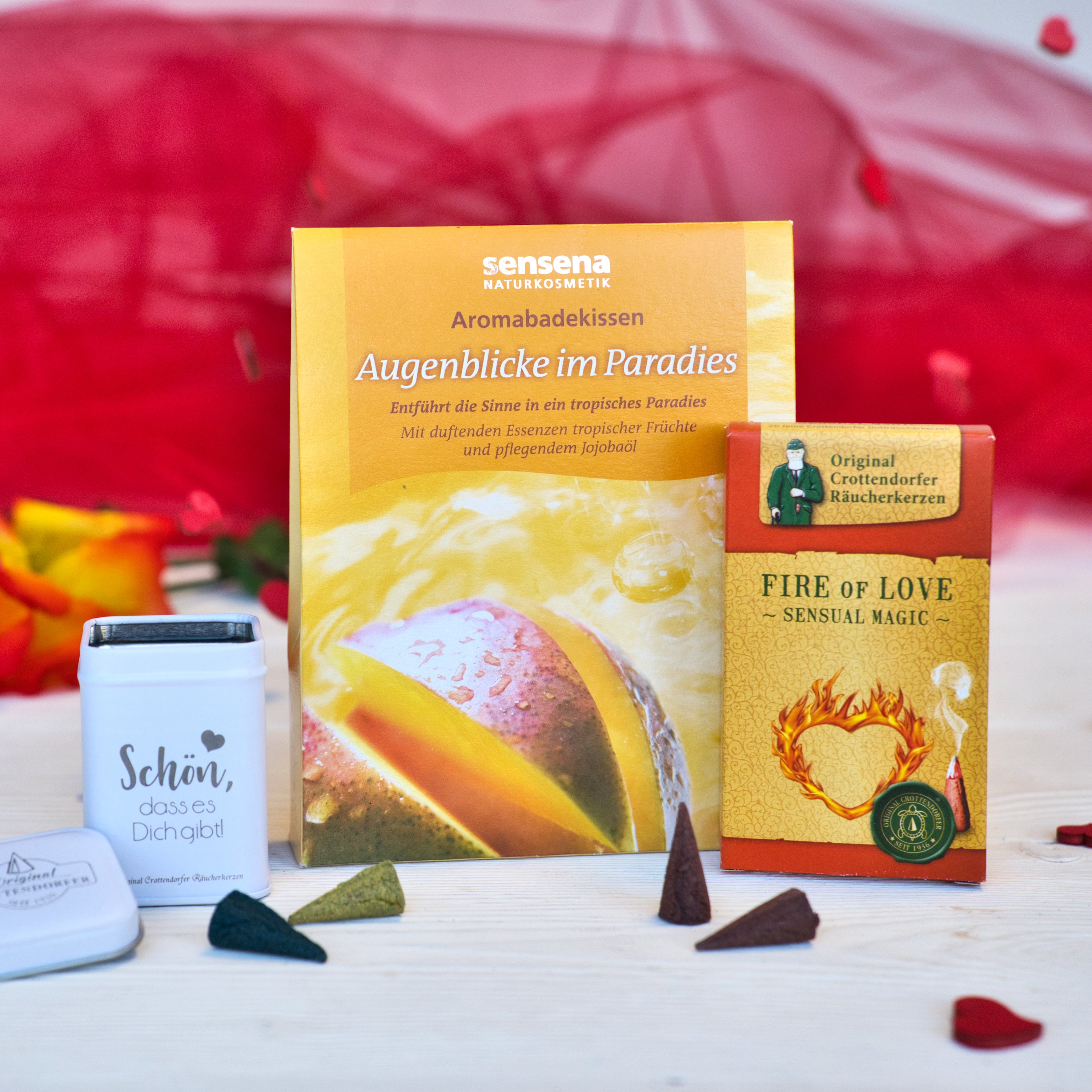 Drei Wellnessprodukte aus dem Geschenkset „Augenblicke der Liebe“ von Original Crottendorfer liegen auf einer hellen Fläche, im Hintergrund sind rote Blütenblätter und ein rotes Tuch zu sehen.