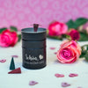 Eine schwarze Räucherfass Sprüche von Original Crottendorfer Räucherkerzen ist auf einem weißen Untergrund mit rosa Rosen und Herzen platziert. Die Szenerie wird durch einen rosa Hintergrund mit Herzmuster aufgewertet.