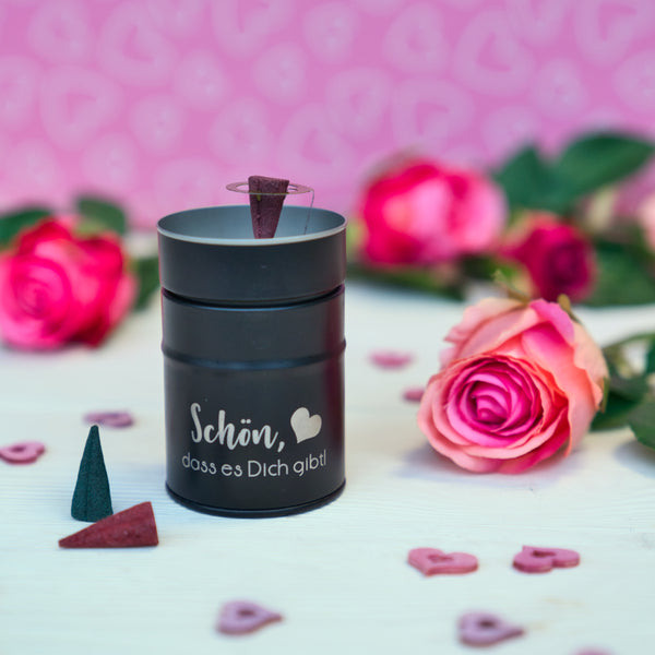 Eine schwarze Räucherfass Sprüche von Original Crottendorfer Räucherkerzen ist auf einem weißen Untergrund mit rosa Rosen und Herzen platziert. Die Szenerie wird durch einen rosa Hintergrund mit Herzmuster aufgewertet.