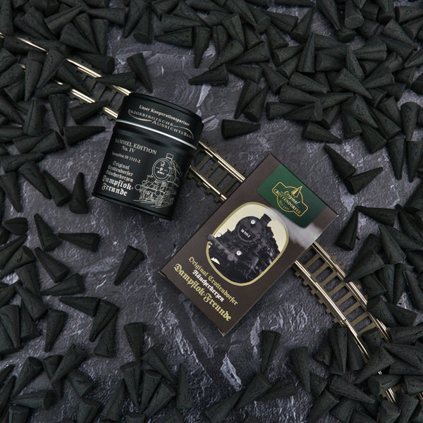 Ein Satz dunkler Räucherkerzen-Kegel liegt auf einer strukturierten Oberfläche neben einem Metallgleis, daneben ein schwarzer Behälter und eine Schachtel mit einem Dampflok-Bild. Beide mit der Aufschrift „Räucherfass – Sammel Edition No. IV Dampflok-Freunde“ von Original Crottendorfer Räucherkerzen versehen, was das Räuchervergnügen-Erlebnis noch verstärkt.