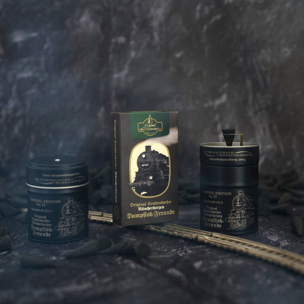 Zwei schwarze Dosen mit Vintage-Etiketten flankieren eine Schachtel mit Dampflok-Illustration im Räucherfass – Sammel Edition Nr. IV Dampflok-Freunde von Original Crottendorfer Räucherkerzen, mit kleinen schwarzen Kohlestücken und Spielzeugeisenbahn-Gleisstücken, die nostalgische Räuchervergnügen auf dunklem, rauchigem Hintergrund erzeugen.