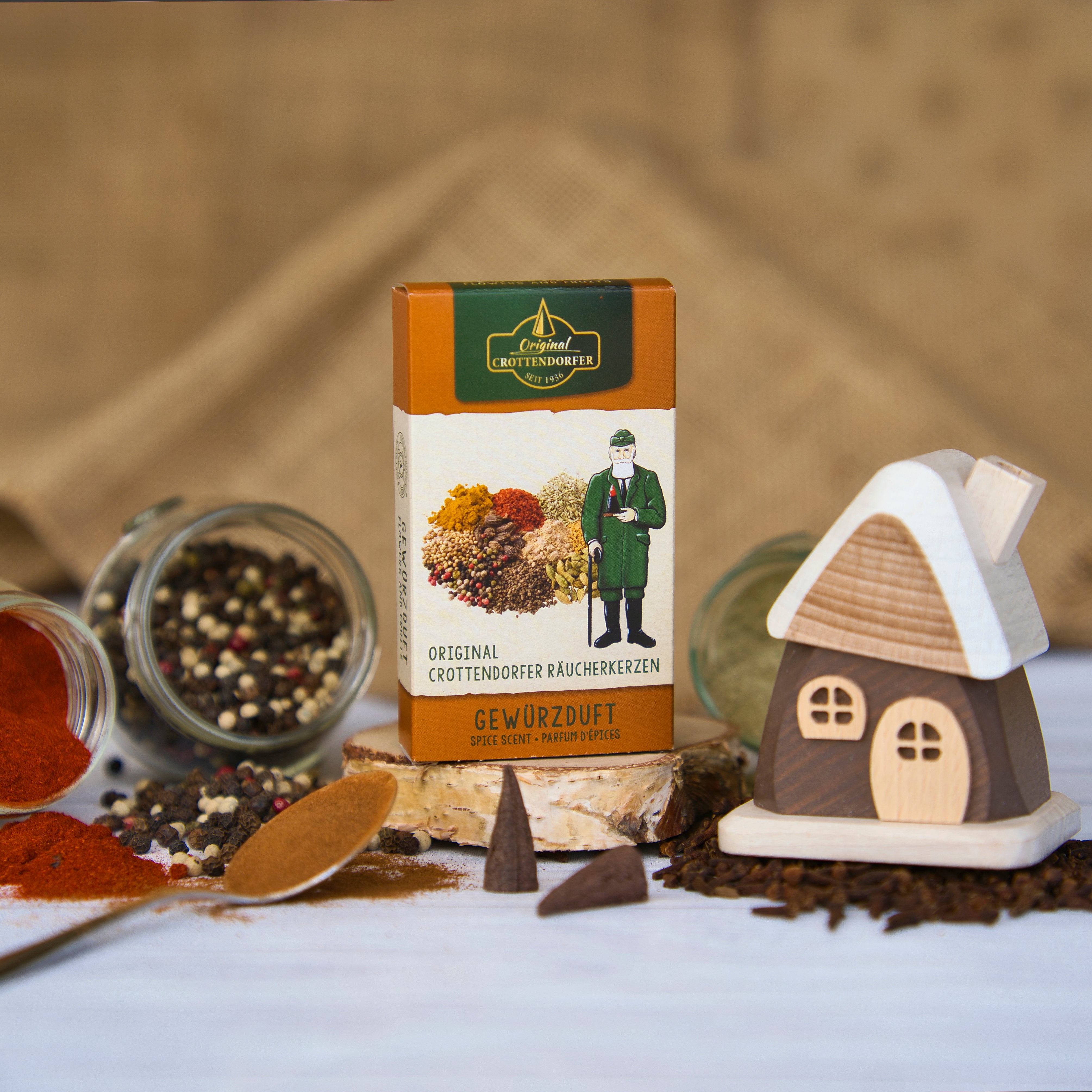 Eine Schachtel Räucherkegel mit bunten Gewürzen und der Abbildung eines Trachtenmannes steht neben einem bezaubernden Räucherhäuschen aus braunem Holz von Holzwaren Egermann aus dem Erzgebirge, ergänzt durch offene Gläser mit losen Gewürzen und Pfefferkörnern auf einer weißen Oberfläche.
