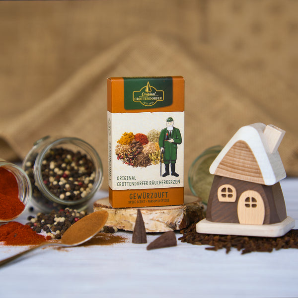 Eine Schachtel Räucherkegel mit bunten Gewürzen und der Abbildung eines Trachtenmannes steht neben einem bezaubernden Räucherhäuschen aus braunem Holz von Holzwaren Egermann aus dem Erzgebirge, ergänzt durch offene Gläser mit losen Gewürzen und Pfefferkörnern auf einer weißen Oberfläche.