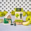 Ein bezauberndes Räucherhäuschen aus Holz grün von Holzwaren Egermann und eine Schachtel mit Räucherkegeln mit grünem Apfelduft sitzen anmutig auf einer Holzscheibe. Mit Äpfeln und Zapfen vor einer weißen Wand erinnert die Schachtel mit ihrem Apfelbild und der traditionellen Figur an die handwerkliche Note der erzgebirgischen Handarbeit.
