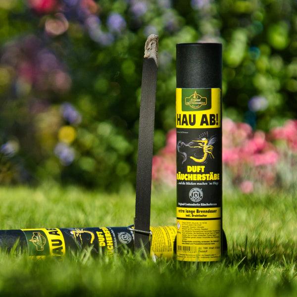 Ein schwarzer HAU AB! Dufträucherstäbe von Original Crottendorfer Räucherkerzen steht im Gras neben seiner gelb-schwarzen Röhre mit der Aufschrift Mückenschutz und Insektenmotiven vor einem unscharfen Gartenhintergrund.