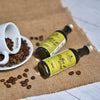 Zwei Flaschen Original Crottendorfer Hausgemachter Kaffeesirup 50 ml stehen auf Sackleinen. Daneben verschütten eine weiße Tasse und Untertasse Kaffeebohnen auf einer weißen Holzoberfläche. Die Flaschen mit den gelben Etiketten eignen sich ideal für die Zubereitung von Eiskaffee oder einzigartigen Cocktails.