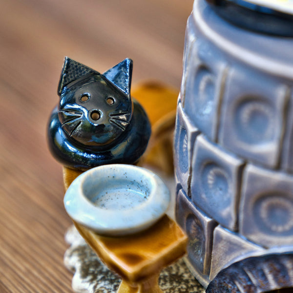 Eine Nahaufnahme einer schwarzen Katzenfigur aus Keramik mit weißen Schnurrhaaren sitzt neben einer leeren Schale auf einer Holzfläche neben einem strukturierten Keramik-Räucherofen Mini rund, grau der Keramikwerkstatt Carola Busch.