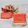 Ein Stück rosa Schafmilchseife Wassermelone von Ovis mit Schaf-Logo und der Aufschrift „SCHAFMILCHSEIFE“ liegt auf einem kleinen runden Holzpodest und verströmt natürliche Frische. Im unscharfen Hintergrund runden weitere Seifenstücke sowie dekorative Blumen und Blätter die ruhige Szene ab.