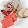 Ovis Schafmilchseife Wassermelonen-Riegel liegen auf einer weichen, weißen Oberfläche, begleitet von Birkenholzscheiben und einem Zweig. Ein Strauß rosa und lila Blumen bereichert den Hintergrund und verleiht dem rustikalen Ambiente natürliche Frische.