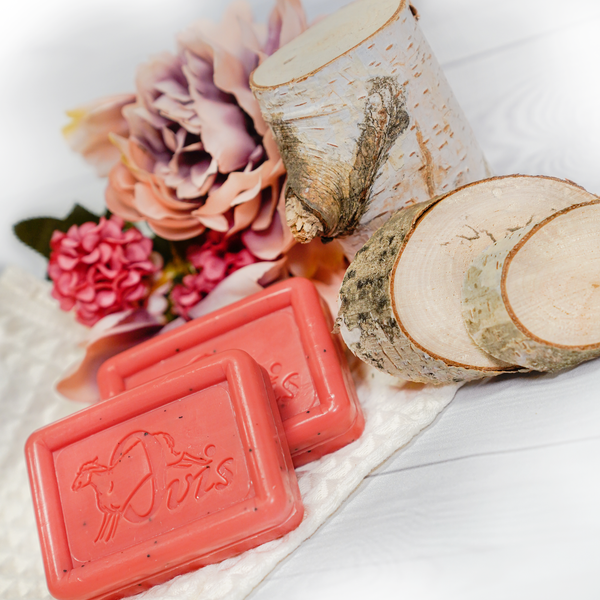 Ovis Schafmilchseife Wassermelonen-Riegel liegen auf einer weichen, weißen Oberfläche, begleitet von Birkenholzscheiben und einem Zweig. Ein Strauß rosa und lila Blumen bereichert den Hintergrund und verleiht dem rustikalen Ambiente natürliche Frische.