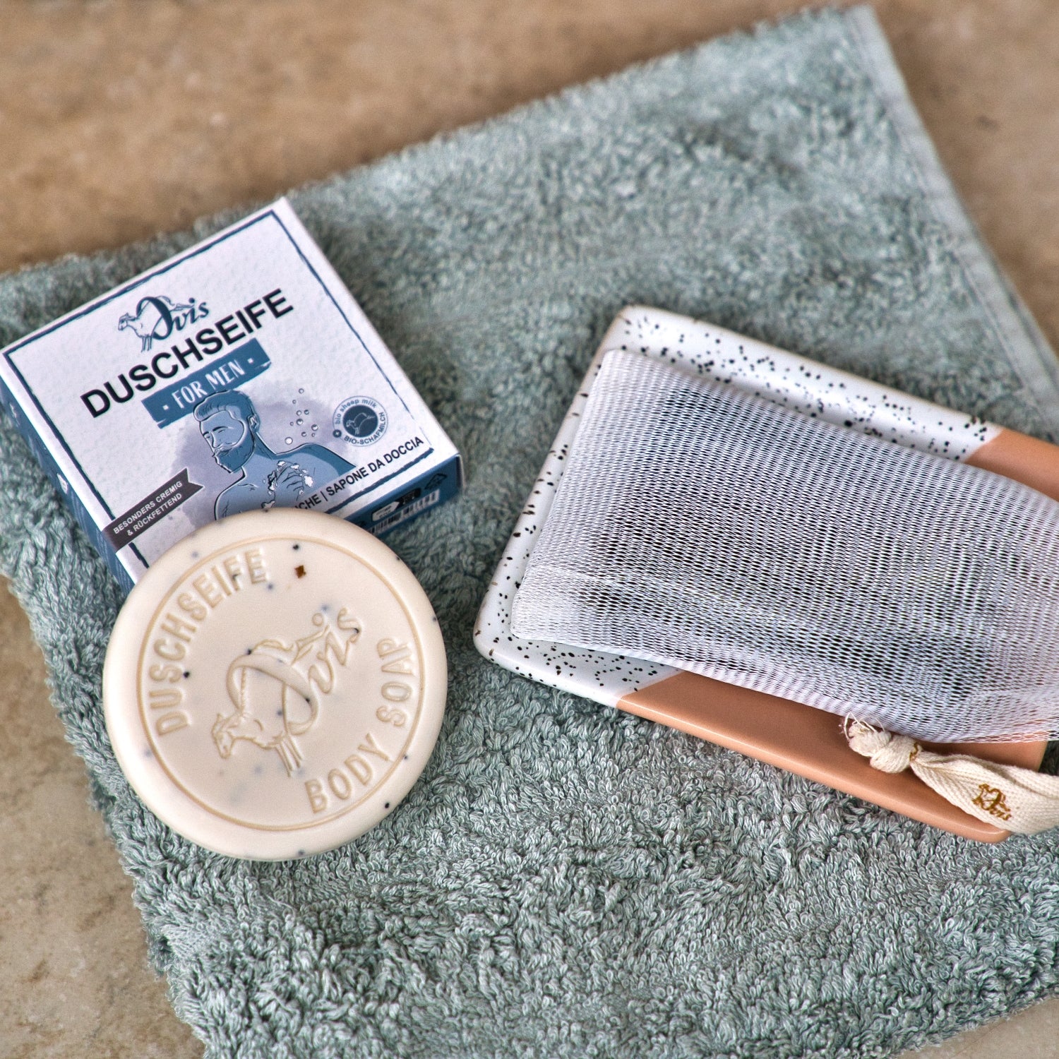 Eine runde Ovis Seife, eine Duschseife, der Ovis Seifenschaumbeutel aus Maisstärke und ein gefaltetes grünes Handtuch sind auf einer beigen Fläche angeordnet - ideal für ein umweltfreundliches, biologisch abbaubares Badezimmer-Set.
