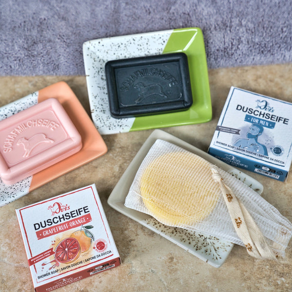 Vier Ovis-Seifen ruhen auf der Porzellanseifenschale eckig, fleckig: rosa, schwarze und gelbe Stangen - eine davon in einem Netzbeutel - und zwei Duschseifenboxen auf Badezimmerfliesen mit einem lila Handtuch im Hintergrund sorgen für ein echtes Design-Highlight.
