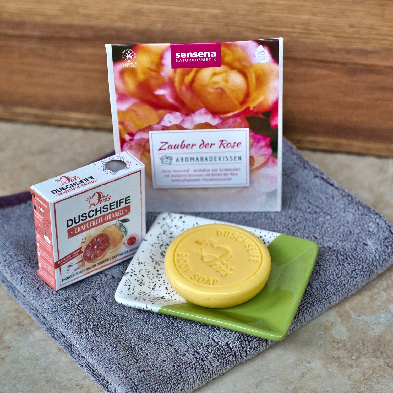 Eine Ovis Porzellanseifenschale eckig, fleckig hält eine grapefruit-orange Seife neben ihrer Schachtel und ein nach Rosen duftendes Badekissenpaket, alles auf einem gefalteten grauen Handtuch platziert - ein Design-Highlight für jedes stilvolle Badezimmer.