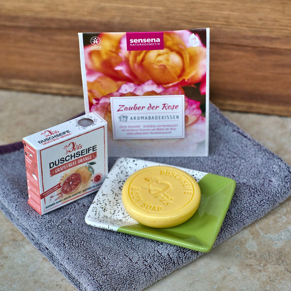 Eine Ovis Porzellanseifenschale eckig, fleckig hält eine grapefruit-orange Seife neben ihrer Schachtel und ein nach Rosen duftendes Badekissenpaket, alles auf einem gefalteten grauen Handtuch platziert - ein Design-Highlight für jedes stilvolle Badezimmer.