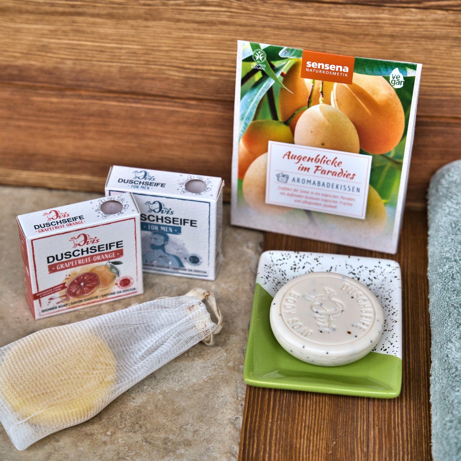 Auf einer Steinfläche befinden sich Ovis-Seifenschaumbeutel, zwei kleine Schachteln mit Ovis-Seife, ein Netzbeutel mit runder Ovis-Seife, eine runde Seife auf einer Schale und ein mit Mango-Motiven verzierter Beutel.