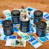 Fünf schwarze "Räucherfass runder Geburtstag"-Dosen von Original Crottendorfer Räucherkerzen sind im Kreis auf Holz angeordnet, umgeben von buntem Spielgeld. Die mittlere Dose ist offen mit einem roten Zeiger - ideal für Räuchervergnügen to go!.