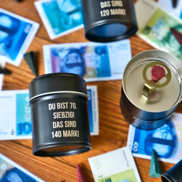 Mehrere schwarze Blechdosen von Original Crottendorfer Räucherkerzen, beschriftet mit "Räucherfass runder Geburtstag", liegen auf einem Holztisch mit buntem Spielgeld. Eine geöffnete Dose zeigt einen Münzschlitz und ein Wachssiegel - perfekt für das Räuchervergnügen unterwegs.