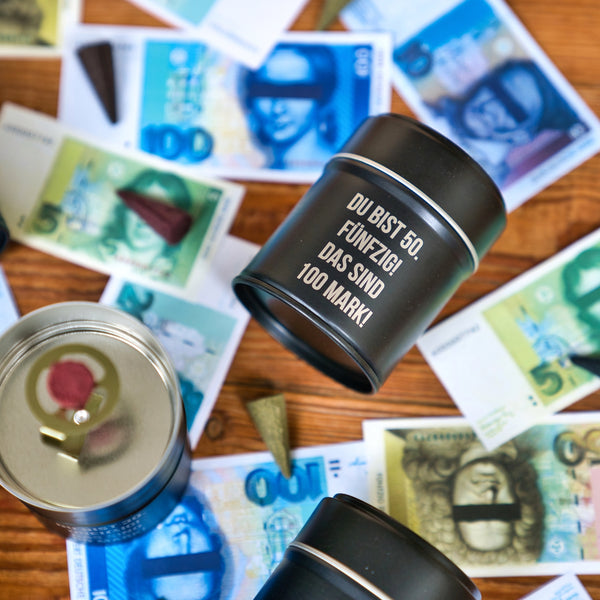 Drei schwarze Original Crottendorfer Räucherkerzen Räucherfass runder Geburtstag Dosen liegen auf Holz inmitten von bunten Geldscheinen. Auf der mittleren Dose steht: Du bist 50. Fünfzig! Das sind 100 Mark!" - ideal für das Räuchervergnügen unterwegs.