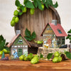 Zwei dekorative Räucherhäuschen-Brauhaus-Modelle stehen auf Holz, daneben ein Räucherkerzenhalter. Frischer Hopfen, grüne Blätter und ein Holzfass bereichern die Szenerie und sorgen mit Produkten der Original Crottendorfer Räucherkerzen für eine gemütliche, rustikale Atmosphäre.