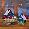 Zwei kleine dekorative Häuser stehen auf Holzflächen mit Tannenzapfen. Ein charmantes Metallhäuschen zeigt einen Schneemann und Tiere, während das andere, genannt Räucherhäuschen Weihnachtsabend von Original Crottendorfer Räucherkerzen, Weihnachtsmann und Rentier vor verschwommenen festlichen Lichtern zeigt.
