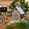 Eine festliche Szene mit dem „Räucherhäuschen Glühweinhütte“ von Original Crottendorfer Räucherkerzen, gestaltet als charmantes Miniaturhaus mit abgebildeten Personen, auf Holz inmitten von Grün und sanft leuchtender Weihnachtsbeleuchtung im Hintergrund.