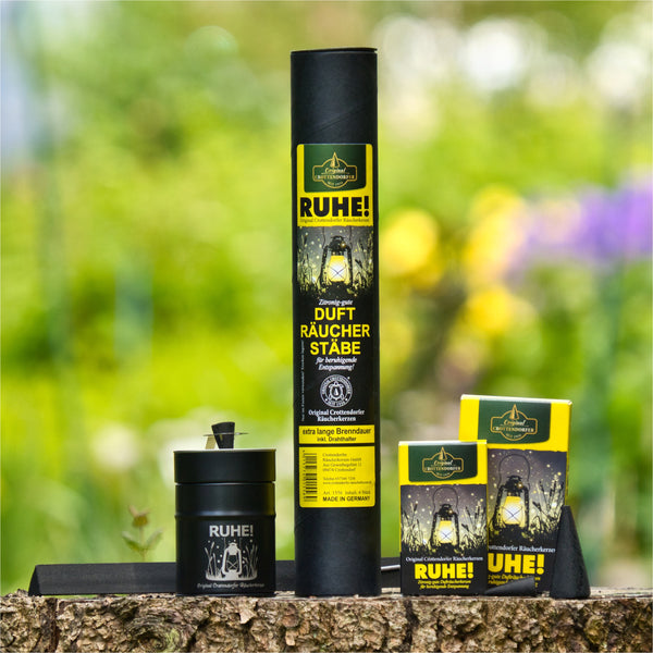 Die „RUHE!“-Kollektion von Original Crottendorfer Räucherkerzen, genannt RUHE! Räucherfass, präsentiert eine hohe zylindrische Verpackung, eine kleine Schachtel, ein Gefäß und einen eleganten schwarzen Räucherstäbchenhalter auf einer Holzfläche im Freien. Die Präsentation lädt zum Räuchervergnügen vor einem verschwommenen Naturhintergrund ein.
