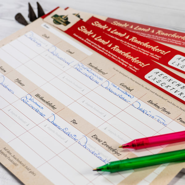 Die Nahaufnahme eines deutschen Dokuments offenbart kreatives Denken mit handgeschriebenem Text in blauer Tinte, roten und beigen Abschnitten sowie gedruckten Überschriften. Zwei Stifte, einer rot und einer grün, liegen auf dem Papier und erinnern an Original Crottendorfers Familienspaß „Stadt-Land-Raacherkerzl“ inmitten teilweise sichtbarer Werkzeuge.