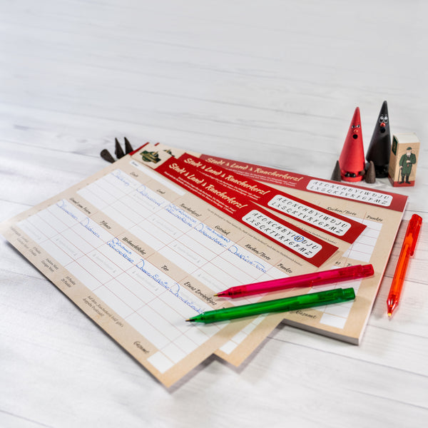 Das Weihnachtsmann-Schreibset von Original Crottendorfer mit zwei Blättern mit Namens- und Adressfeldern, drei bunten Stiften, Dekofiguren und Kärtchen auf heller Holzoberfläche weckt mit dem Familienspaß „Stadt-Land-Raacherkerzl“ die Kreativität für den Weihnachtsspaß.