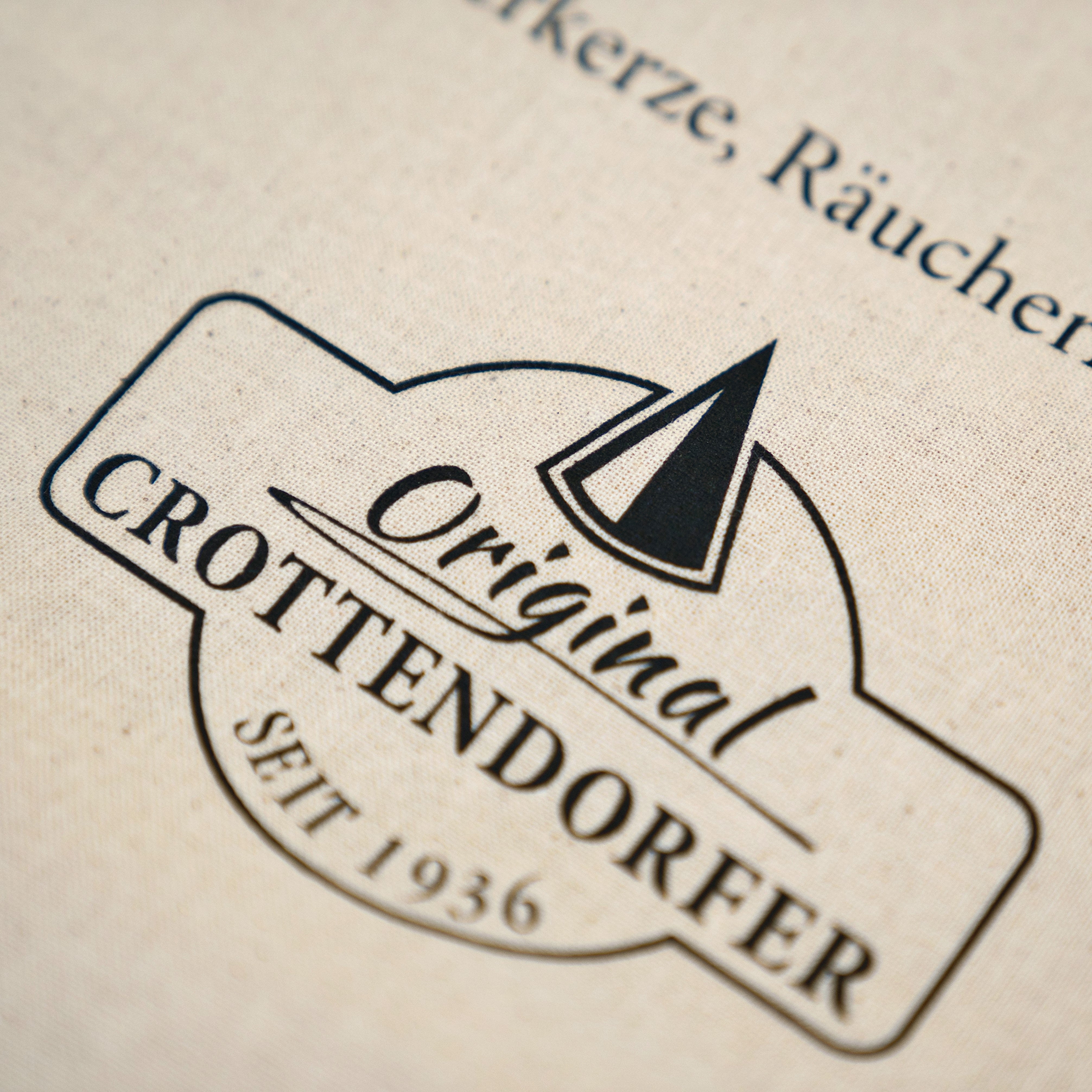 Nahaufnahme eines beigen Stoffetiketts auf dem Stoffbeutel „Raacherkerzl Definition“ oder „Bitte nicht schubsen ...“ von Original Crottendorfer, das das schwarze Logo „Original Crottendorfer Seit 1936“ mit Kegelmotiv und Verweisen auf die erzgebirgische Tradition zeigt.