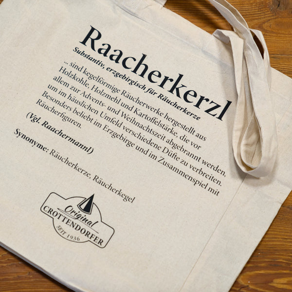 Ein beiger Stoffbeutel von Original Crottendorfer mit dem Aufdruck „Raacherkerzl Definition“ und Hausillustration – perfekt für Fans der Crottendorfer Räucherkerzen und der erzgebirgischen Räucherkultur.