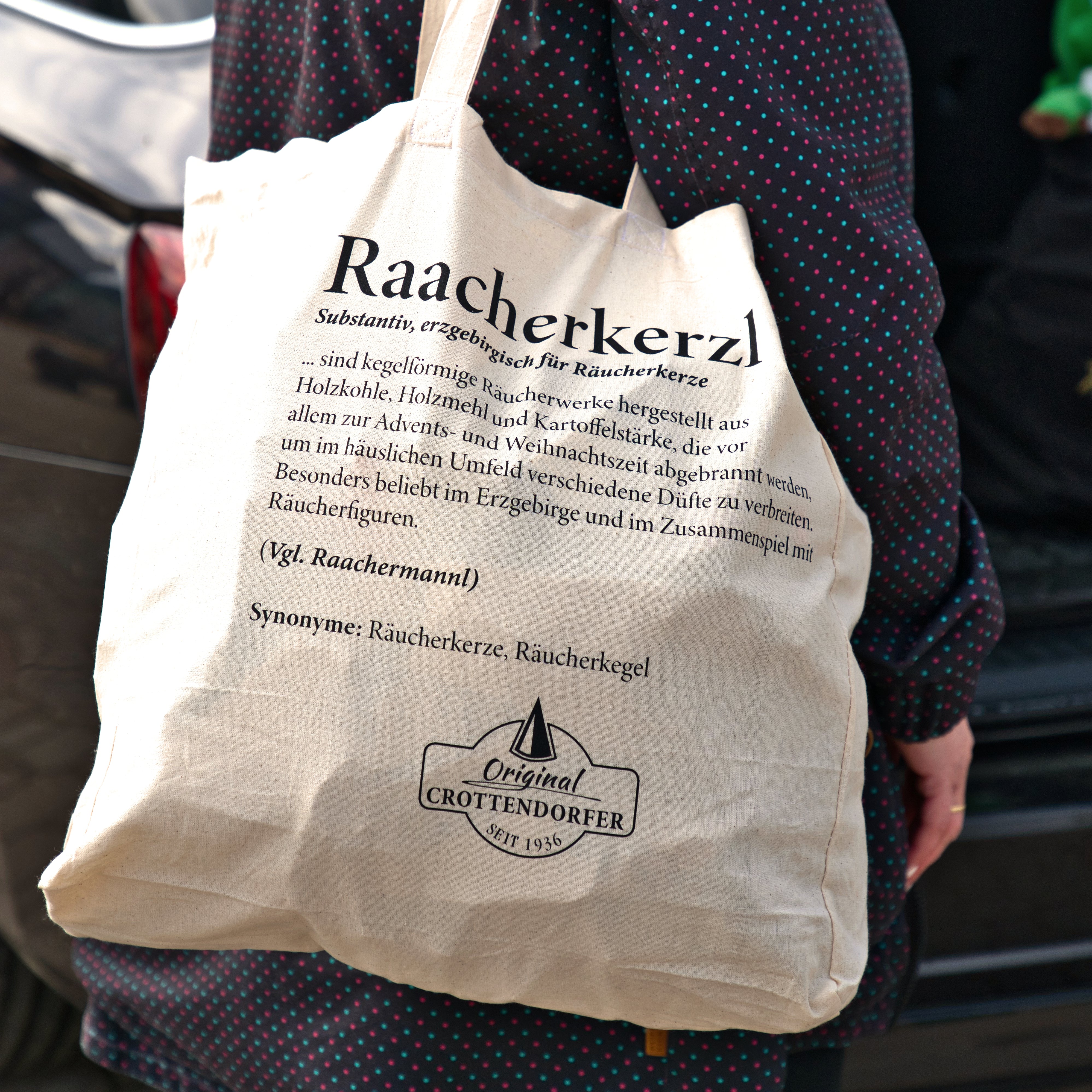 Eine Person trägt einen beigen Stoffbeutel „Raacherkerzl Definition“ oder „Bitte nicht schubsen ...“ von Original Crottendorfer und trägt ein dunkles, gepunktetes Kleid.
