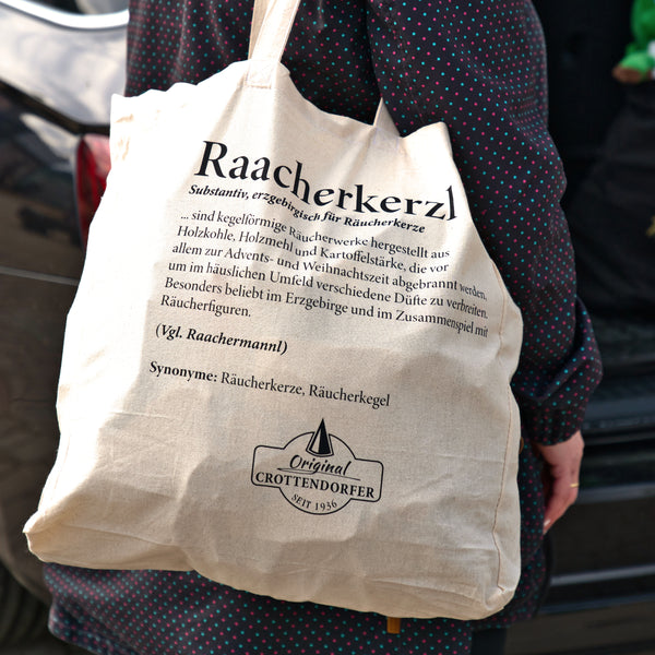 Eine Person trägt einen beigen Stoffbeutel „Raacherkerzl Definition“ oder „Bitte nicht schubsen ...“ von Original Crottendorfer und trägt ein dunkles, gepunktetes Kleid.