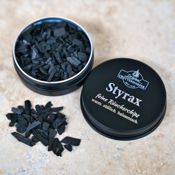 Eine runde Dose mit der Aufschrift "Original Crottendorfer Räucherkerzen - Räucherchips Styrax 10 g in der Schraubdeckeldose" enthält schwarze Räucherchips, von denen einige auf einer beigen Oberfläche verschüttet sind; ideal für sinnliche Duft Räucherwerk-Momente.