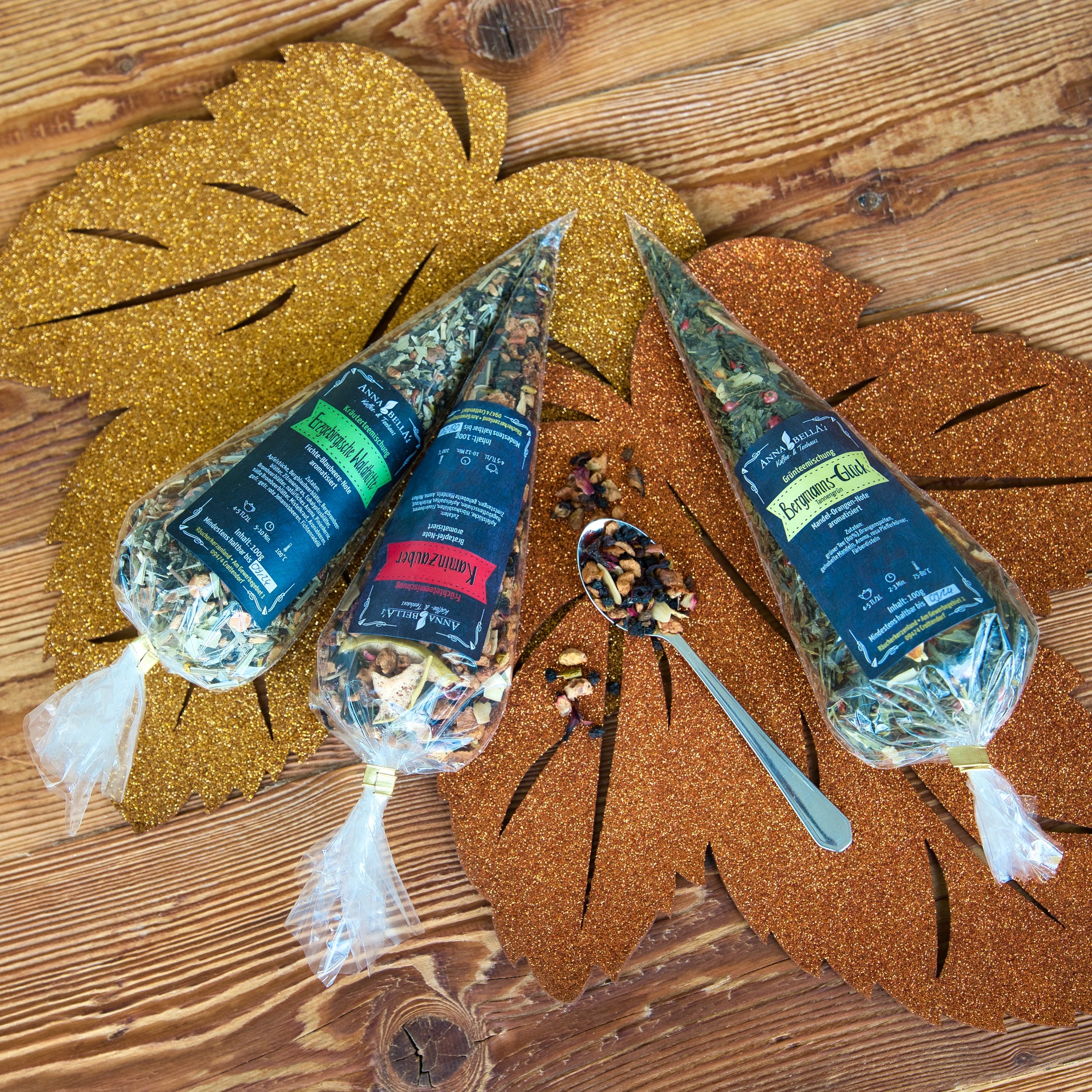 Drei kegelförmige Packungen mit der Original Crottendorfer Kräuterteemischung „Erzgebirgische Waldhütte“ stehen auf glitzernden Blatt-Tischsets, daneben ein silberner Löffel und Teeblätter. Jede Packung hat eine einzigartige Etikettenfarbe, die sie optisch ansprechender macht und auf Fichtennadelöl hinweist.