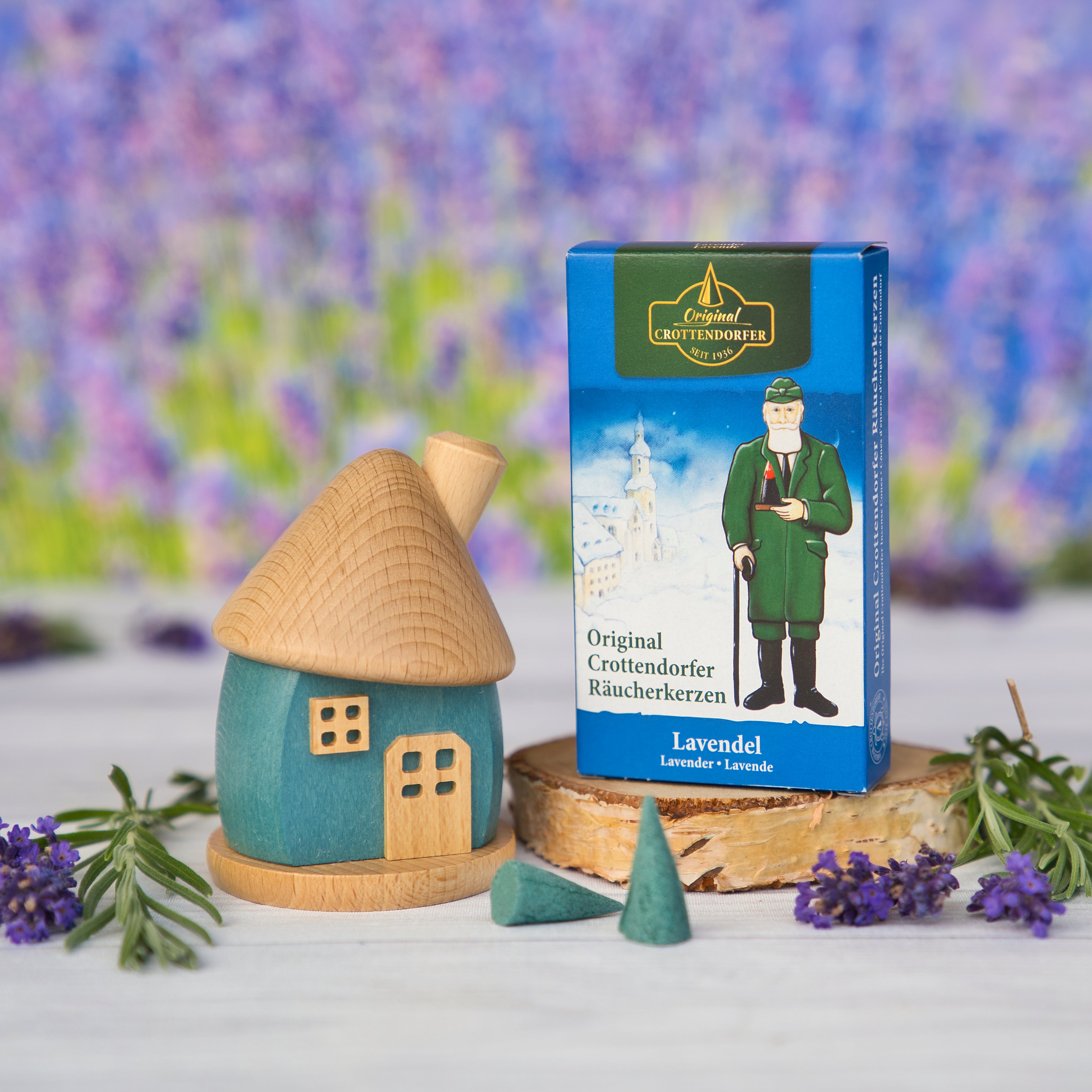 Ein Holzwaren Egermann Räucherhäuschen rund aus Holz blau/natur und eine Schachtel Lavendel-Räucherkegel stehen auf einem Tisch inmitten von Lavendelblüten, im Hintergrund ein verschwommenes Feld. Die Erzgebirgische Handarbeitsbox zeigt eine Figur in traditioneller Kleidung.
